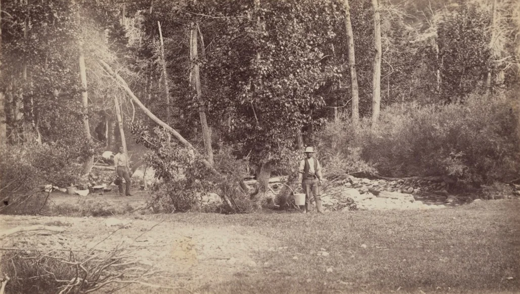 Archival photograph: two men at work beside a creek in open woodland, one standing with a bucket, camp gear on the bank behind them